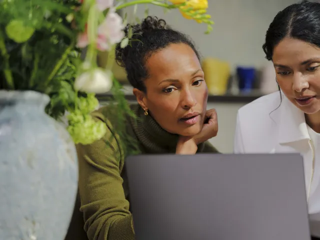 Two women in an office setting look at a laptop screen together, with a potted plant nearby