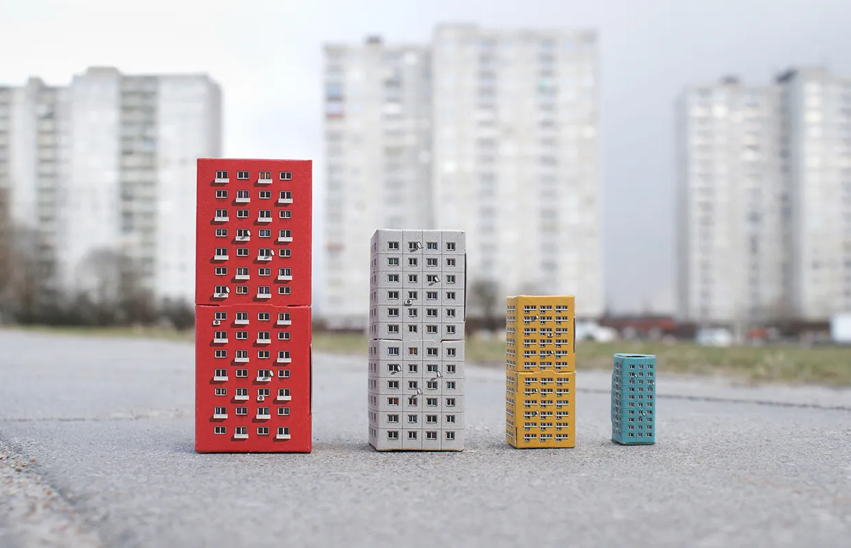Four nested cardboard buildings in red, grey, yellow and turquoise from Blokoshka, arranged in size order on pavement with Soviet-era apartment blocks blurred in background
