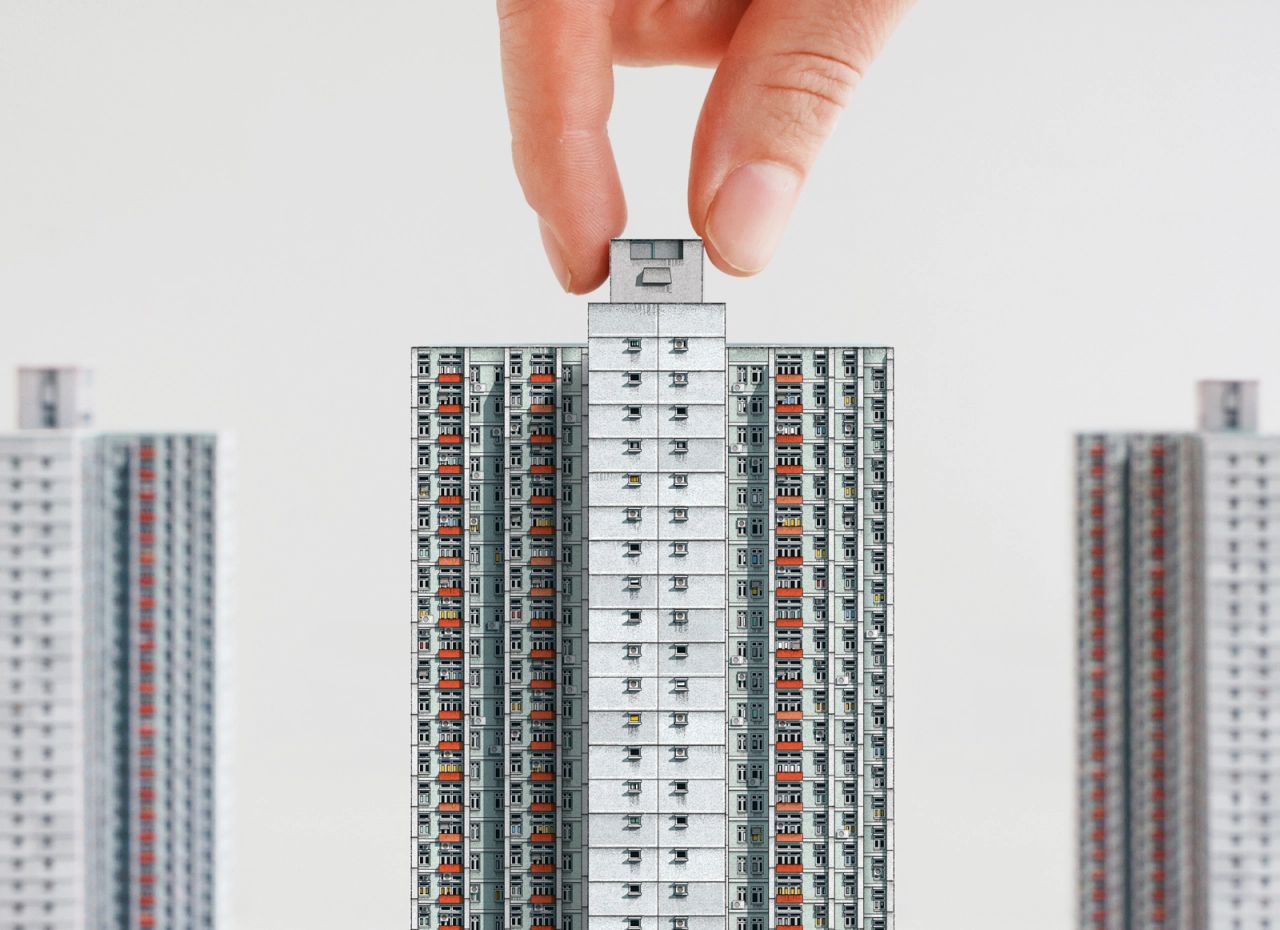 Hand holding a detailed paper model of a Hong Kong public housing tower block, with blurred similar buildings in the background