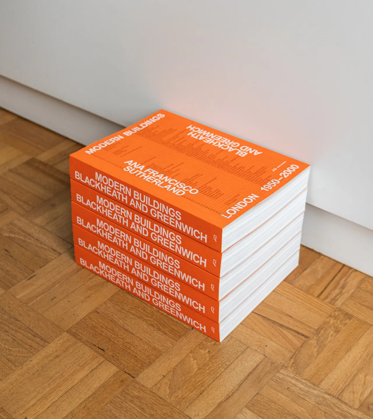 Stack of six copies of Modern Buildings in Blackheath and Greenwich book with bright orange spines on wooden floor against white wall