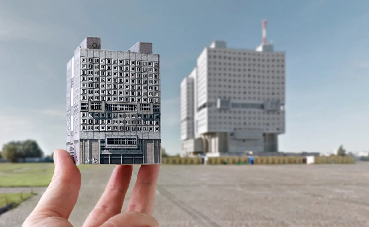 Hand holding a paper model of a modernist building with a large brutalist structure visible in the background across an empty field