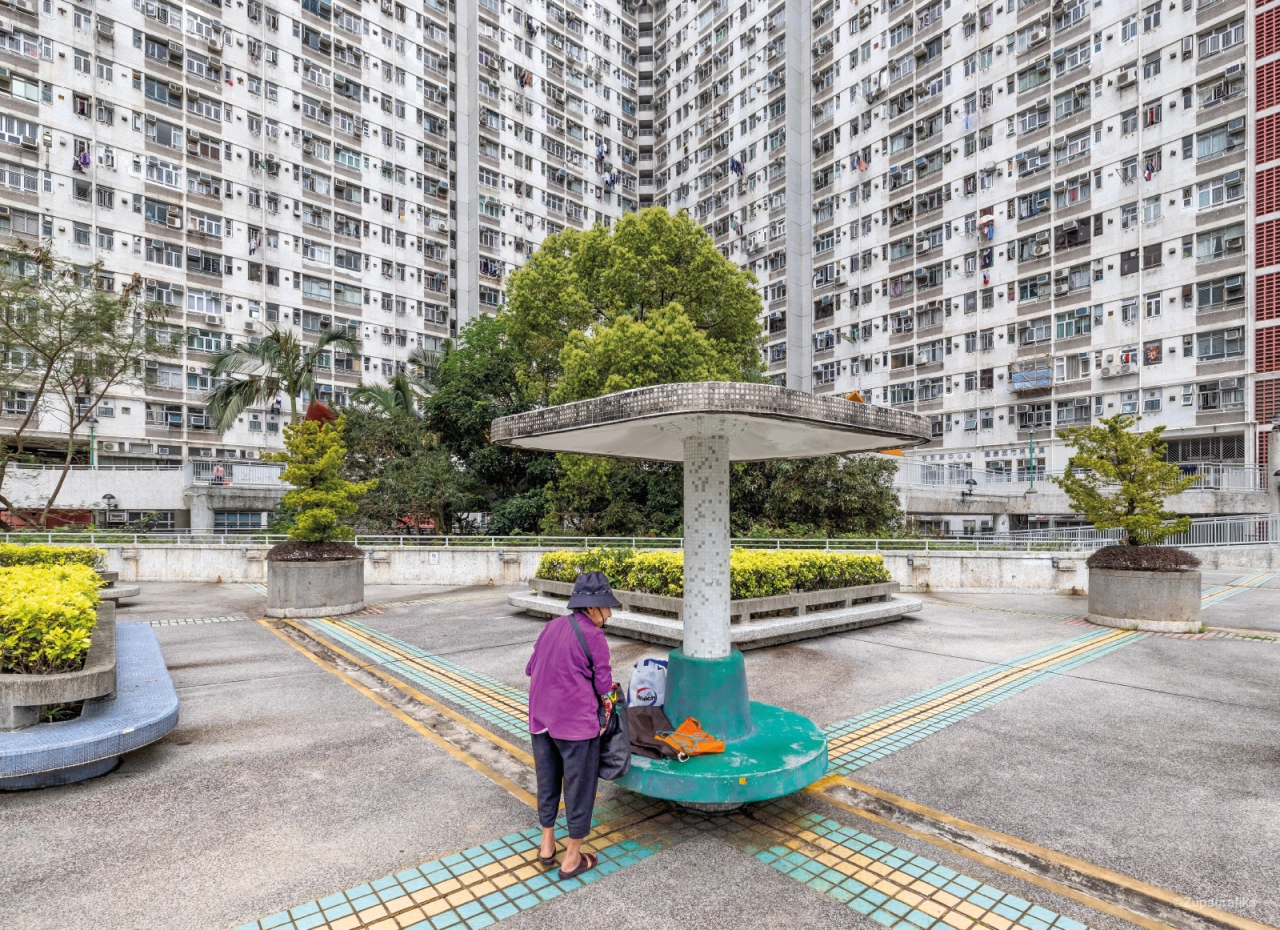Person in pink jacket stands at colourful ground markings beneath a concrete canopy structure in a Hong Kong public housing estate courtyard