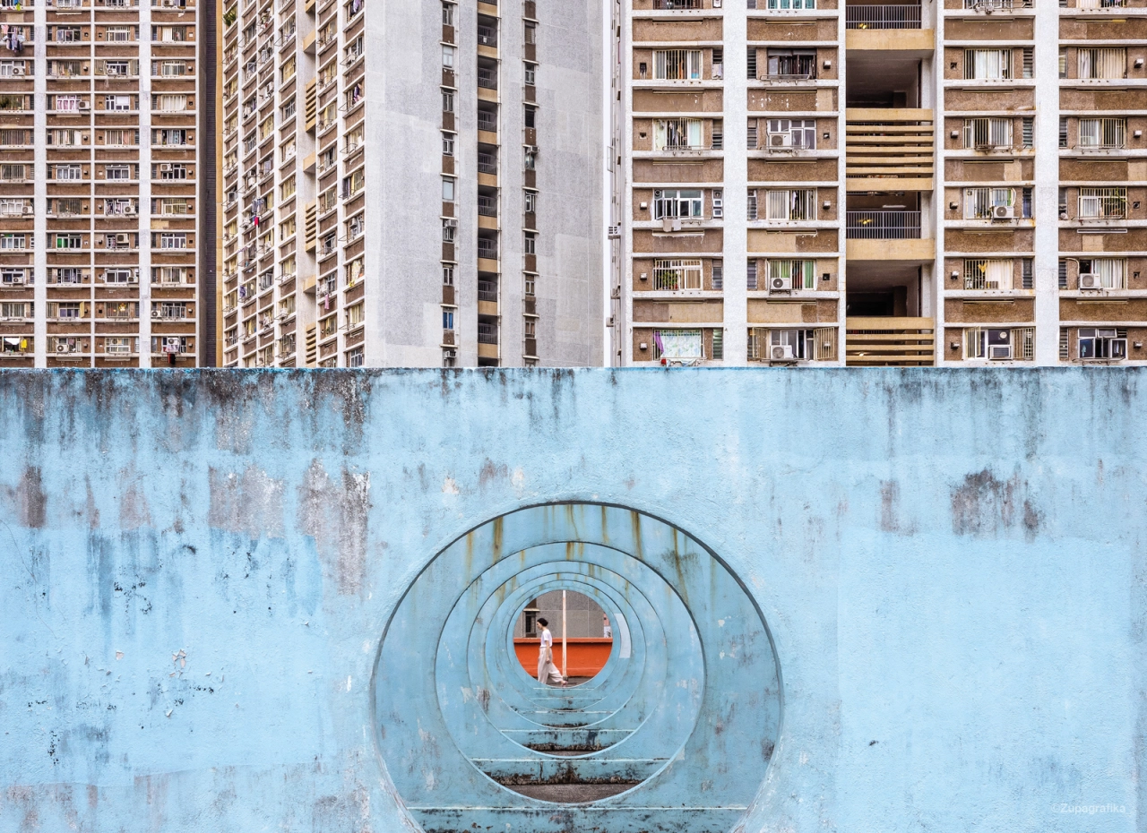 Weathered blue wall with concentric circular metal rings in the centre, with Hong Kong public housing tower blocks visible above