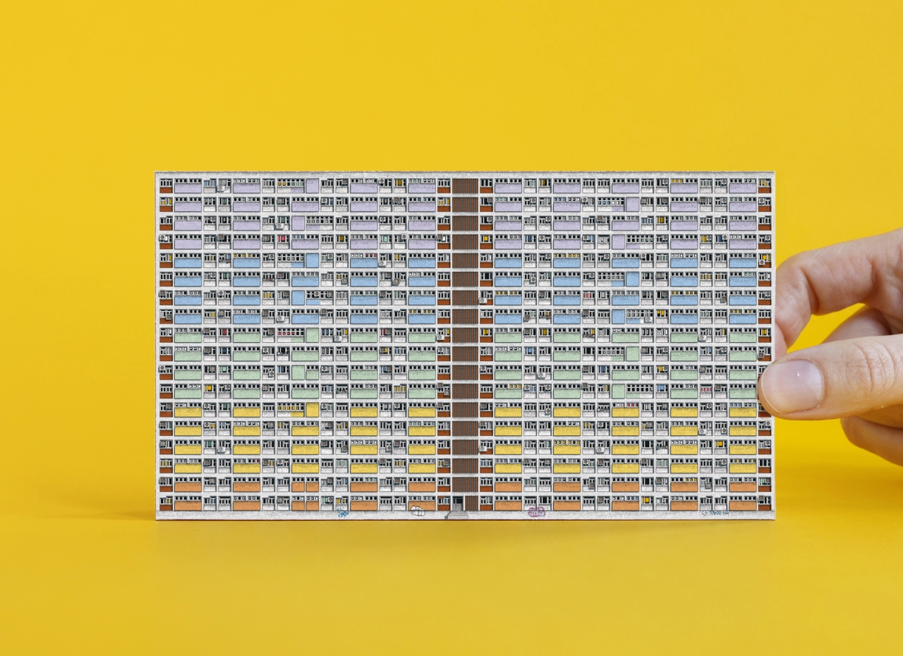 Hand holding a yellow pencil pointing to an intricate foldout paper model of a dense Hong Kong public housing estate against a bright yellow background